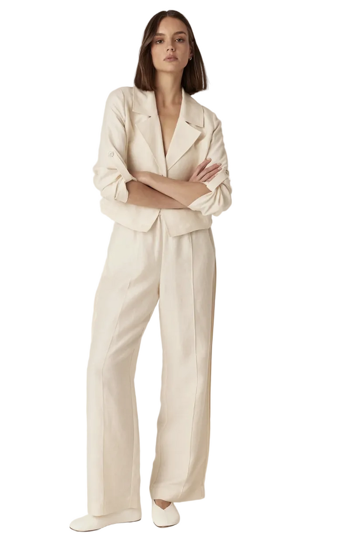 A woman stands against a plain light beige studio background wearing a matching cream-colored tailored set consisting of a cropped blazer and wide-leg trousers, paired with white slip-on shoes.