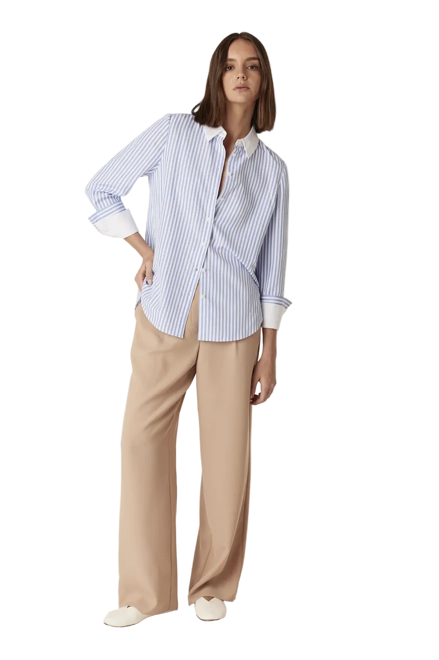 A woman modeling a light blue and white vertically striped button-down shirt with beige wide-leg trousers and white slip-on shoes, photographed against a plain light beige studio background.