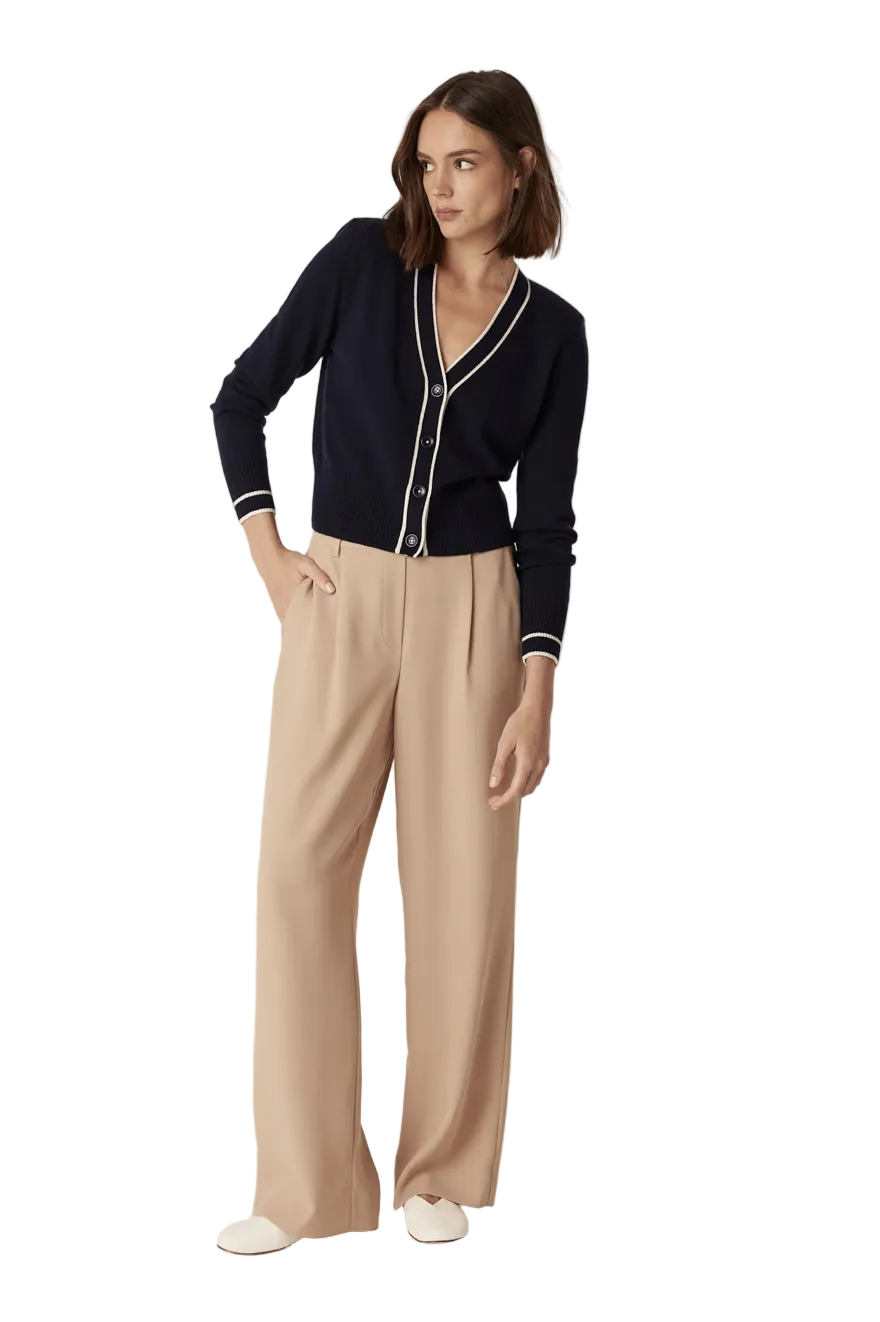 A woman modeling a dark navy blue cardigan with white trim and beige wide-leg trousers against a plain light beige studio background.