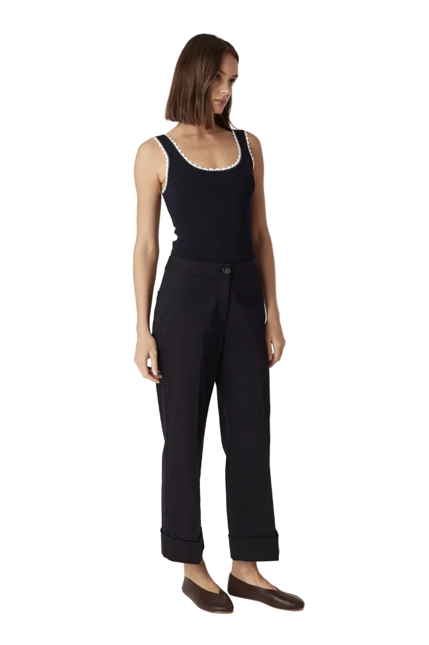 A woman modeling a black sleeveless top with a white scalloped neckline and black trousers, standing against a plain light beige studio background.