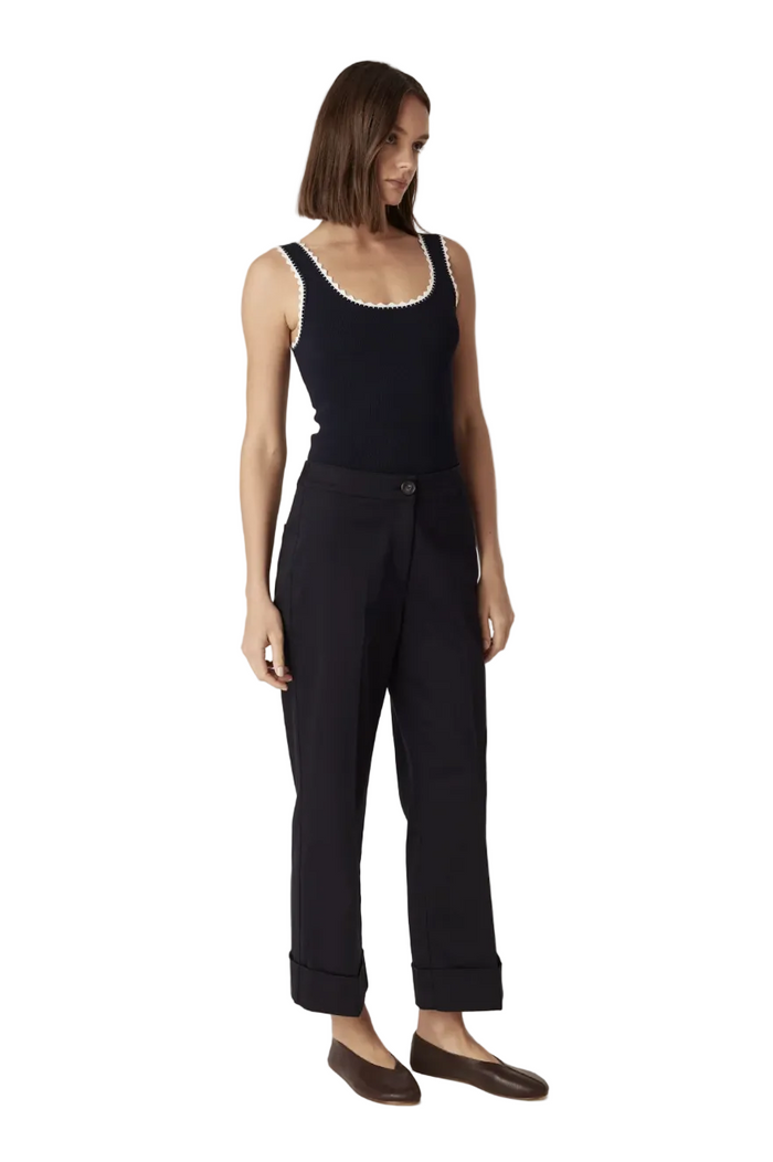 A woman modeling a black sleeveless top with a white scalloped neckline and black trousers, standing against a plain light beige studio background.