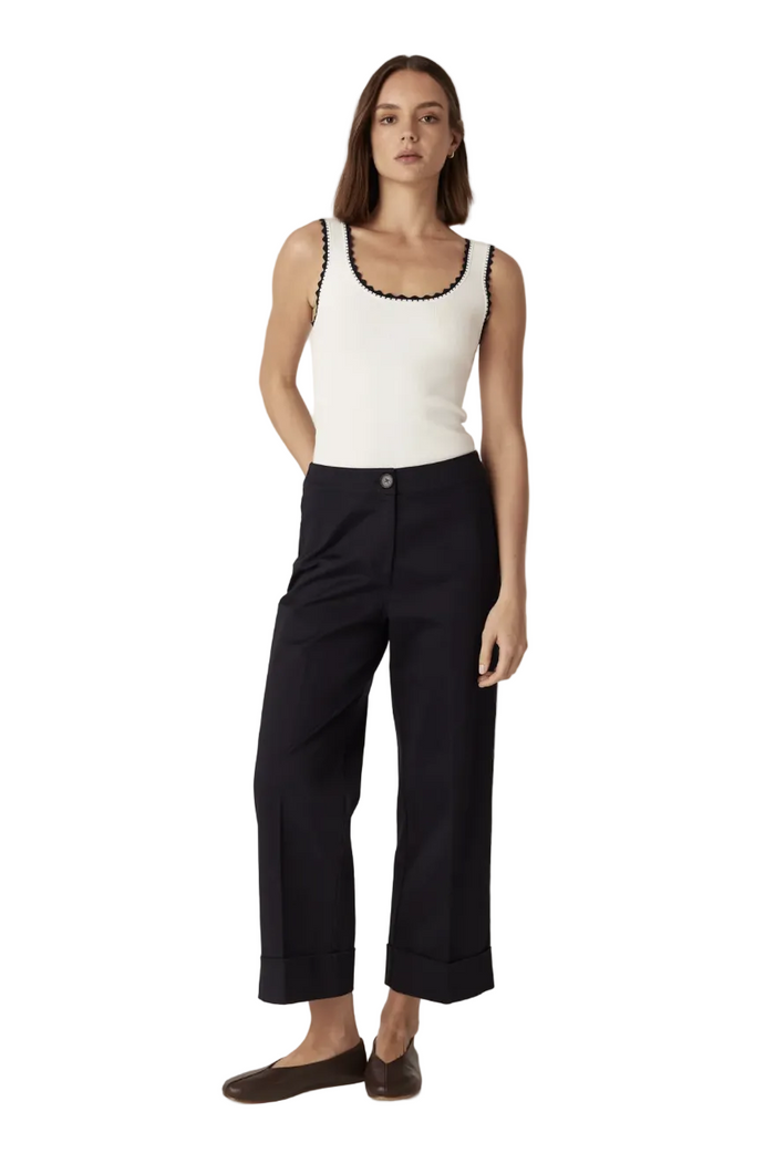 A woman modeling a white ribbed tank top with black trim along the scoop neckline and straps, paired with dark wide-leg cropped trousers and dark flat shoes, against a plain light beige studio background.