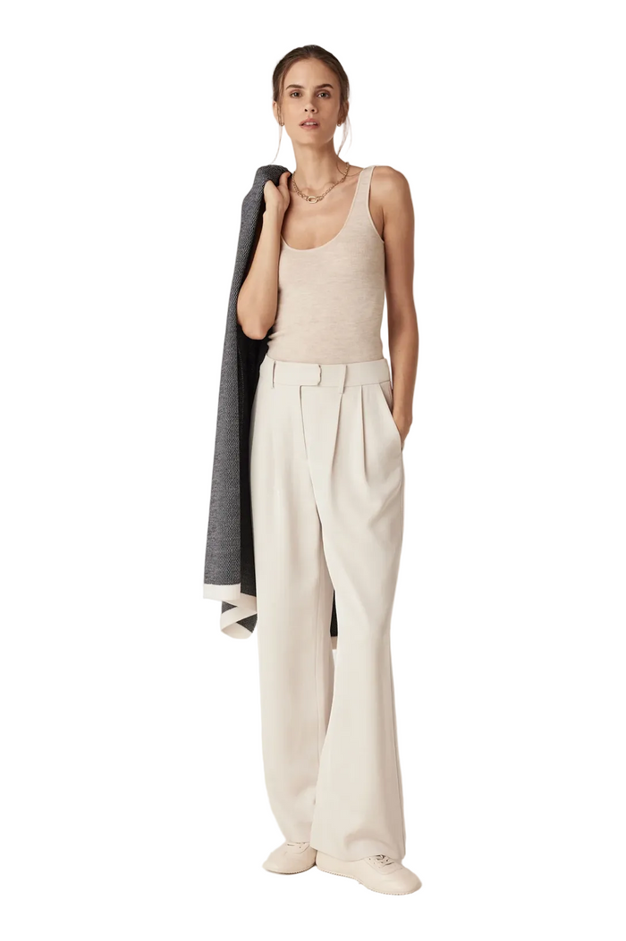 Woman wearing a beige tank top and white pants against a plain background