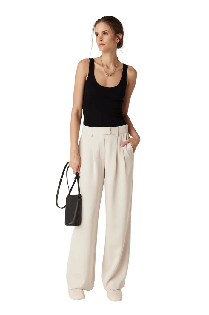 A woman modeling a black sleeveless tank top and high-waisted, wide-leg cream trousers, holding a small black handbag against a plain light gray studio background.